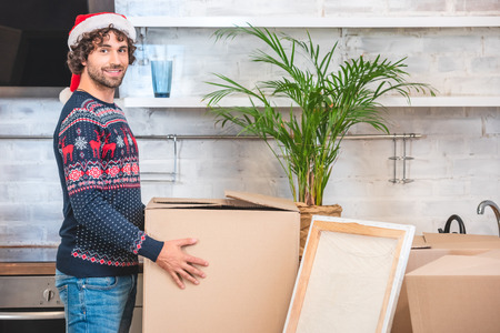 handsome happy young man in santa hat holding cardboard box and smiling at camera during relocation