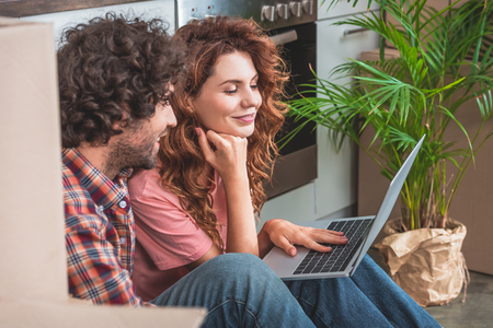 side view of cheerful couple sitting with laptop near cardboard boxes on floor in new kitchenの写真素材