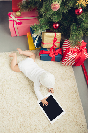 high angle view of little baby with tablet lying on floor near christmas gifts and treeの写真素材