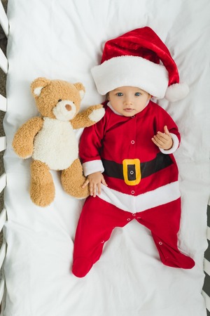 top view of cute little baby in santa suit lying in crib with teddy bear and looking at cameraの写真素材