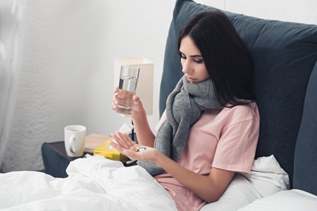 diseased young woman holding pills and glass of water while lying in bedの写真素材