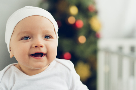 close-up portrait of happy little baby in white hat with blurred christmas tree on backgroundの写真素材