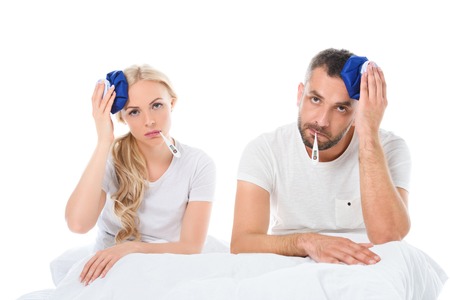 upset couple with electronic thermometers holding ice packs on heads and sitting in bed, isolated on whiteの写真素材