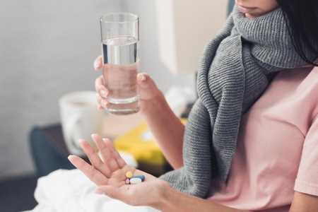 cropped shot of sick young woman holding pills and glass of water while lying in bedの写真素材