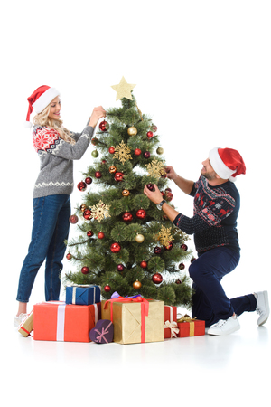 happy couple in santa hats decorating christmas tree with presents, isolated on whiteの写真素材