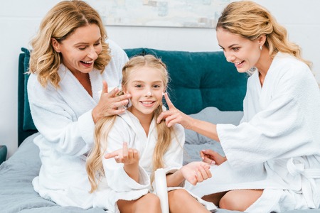 happy child with mother and grandmother in bathrobes sitting on bed at home and applying moisturizing creamの写真素材