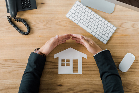 Partial view of businessman covering house paper model with hands at workplace with keyboard and computer mouse, insurance conceptの写真素材