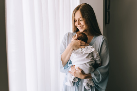 smiling attractive woman carrying little baby boy in front of curtains