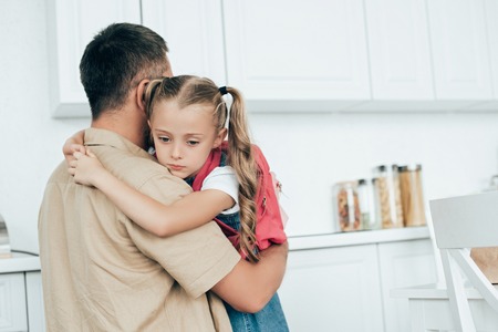 father and sad little daughter with backpack hugging each other in kitchen at home, back to school conceptの写真素材