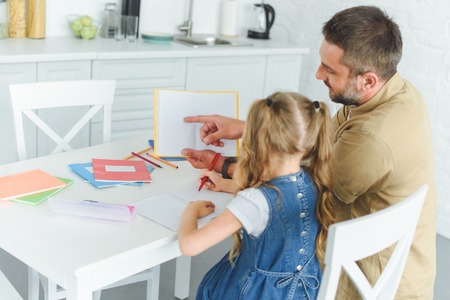 father point at book while doing homework together with daughter in kitchen at home