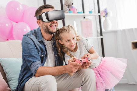 cheerful dad and daughter having fun and playing with joystick and using virtual reality headset on sofaの写真素材