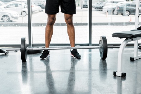 low section of young sportsman standing near barbell in gymの写真素材
