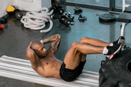 tattooed african american athlete doing abs with legs on tire at gymの写真素材
