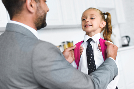 father in suit and little daughter in school uniform with backpack in kitchen at home, back to school conceptの写真素材
