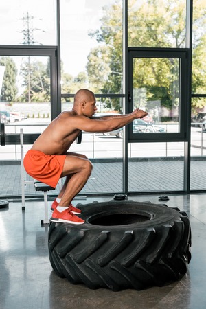 side view of african american athlete doing squats on tire at gymの写真素材