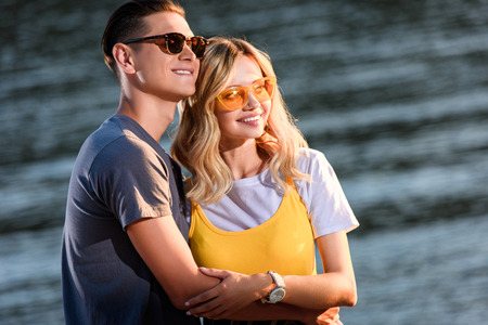 young couple in sunglasses hugging on river beach in evening and looking away