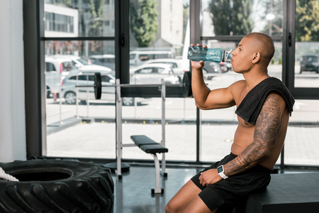 side view of muscular shirtless african american sportsman drinking water in gymの写真素材