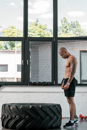 side view of muscular young african american man standing and looking at tire in gymの写真素材