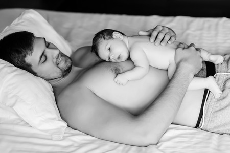 black and white picture of happy shirtless father laying with baby boy in bed at homeの写真素材