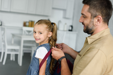 side view of father helping daughter to wear backpack at home, back to school conceptの写真素材