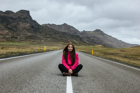 smiling young woman sitting on empty road in iceland with beautiful mountains on backgroundの写真素材