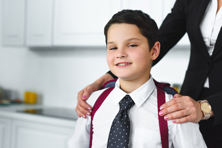 partial view of mother in suit and son in school uniform with backpack in kitchen at home, back to school conceptの写真素材