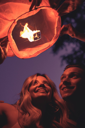 low angle view of smiling young couple launching sky lantern on river beach in evening