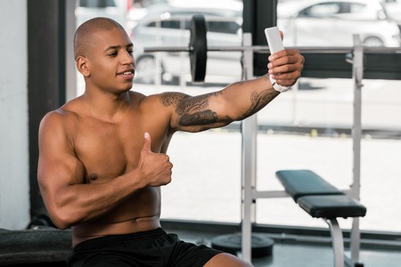 smiling muscular african american man showing thumb up and taking selfie with smartphone in gymの写真素材