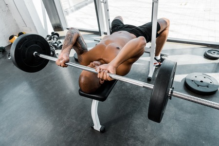 high angle view of muscular african american sportsman lying on bench and lifting barbell in gymの写真素材