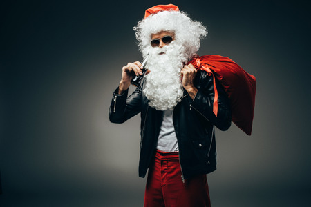 Stylish santa claus in sunglasses and leather jacket drinking cream soda and holding christmas bagの写真素材