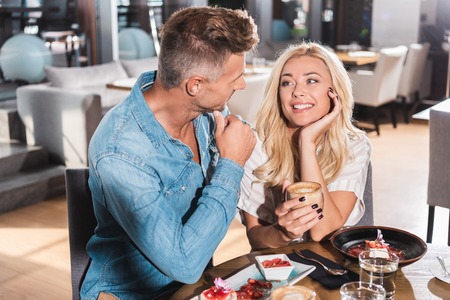 Happy blonde girlfriend holding cup of coffee and looking at boyfriend at table in cafeの写真素材