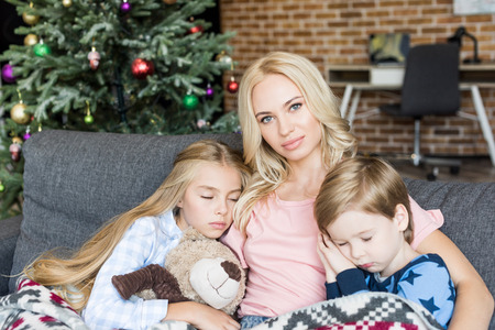 young mother looking at camera while sitting with cute little chikdren in pajamas sleeping on sofa...