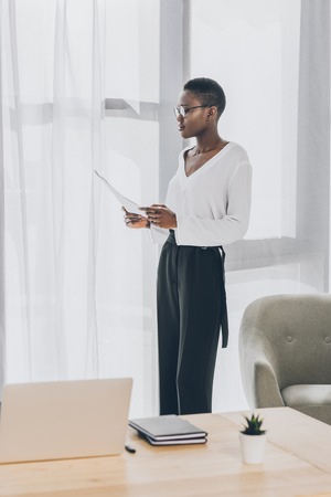 stylish attractive african american businesswoman standing and reading documents in officeの写真素材