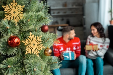 selective focus of decorated christmas tree and couple with gift boxes sitting on sofa behind at homeの写真素材