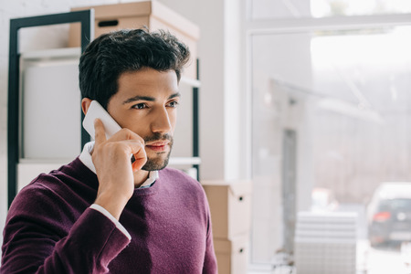 portrait of handsome designer in burgundy sweater talking by smartphone in officeの写真素材