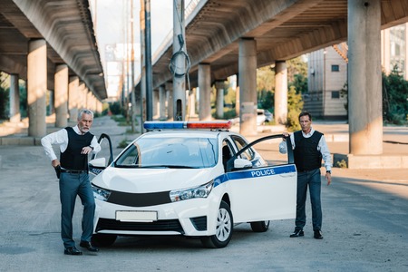 policeman taking off gun from holster and pointing by finger to colleague near car at streetの写真素材