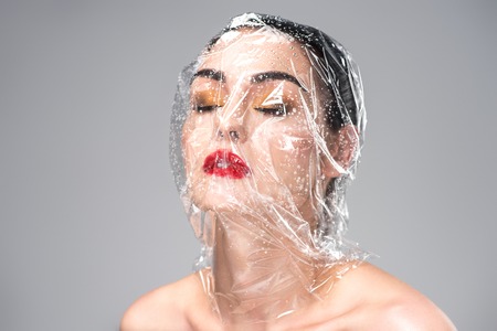 sensual young woman with transparent wet cellophane on head and closed eyes isolated on greyの写真素材