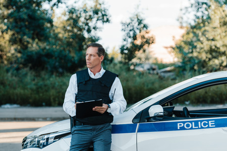 middle aged male police officer in bulletproof vest writing in clipboard near car at streetの写真素材