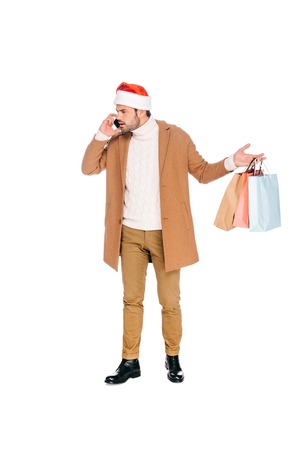 emotional young man in santa hat holding shopping bags and talking by smartphone isolated on whiteの写真素材