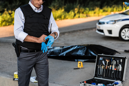 partial view of policeman with gun in holster putting on latex gloves at crime scene with corpse in...