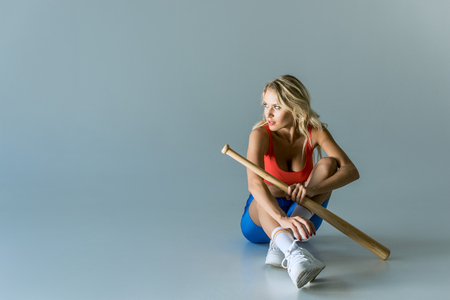 fit young woman with baseball bat sitting on grey and looking awayの写真素材