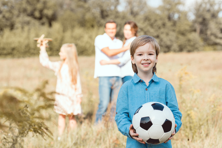 adorable little kid holding ball while his family standing blurred on background in fieldの写真素材