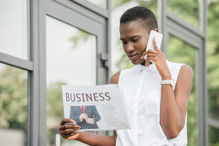 stylish attractive african american businesswoman talking by smartphone and reading newspaper on streetの写真素材