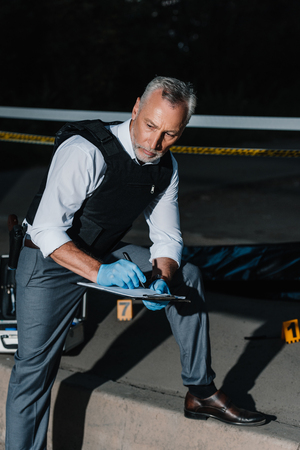 serious middle aged policeman writing in clipboard at crime scene with corpse in body bagの写真素材