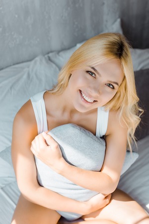 high angle view of happy young woman in underwear sitting with pillow on bed and smiling at cameraの写真素材