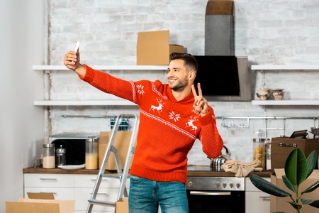 happy young man doing peace sign and taking selfie on smartphone in kitchen with cardboard boxes...