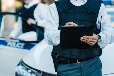 cropped image of male police officer writing in clipboard while his colleague standing near car at streetの写真素材