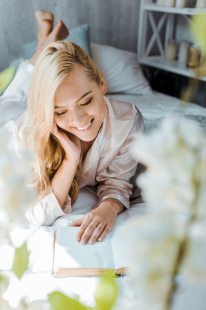 selective focus of beautiful smiling girl in pajamas lying on bed and reading bookの写真素材