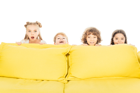 excited and smiling children looking out from yellow sofa and looking at camera isolated on whiteの写真素材