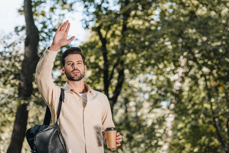 portrait of stylish man with backpack and coffee to go greeting in parkの写真素材